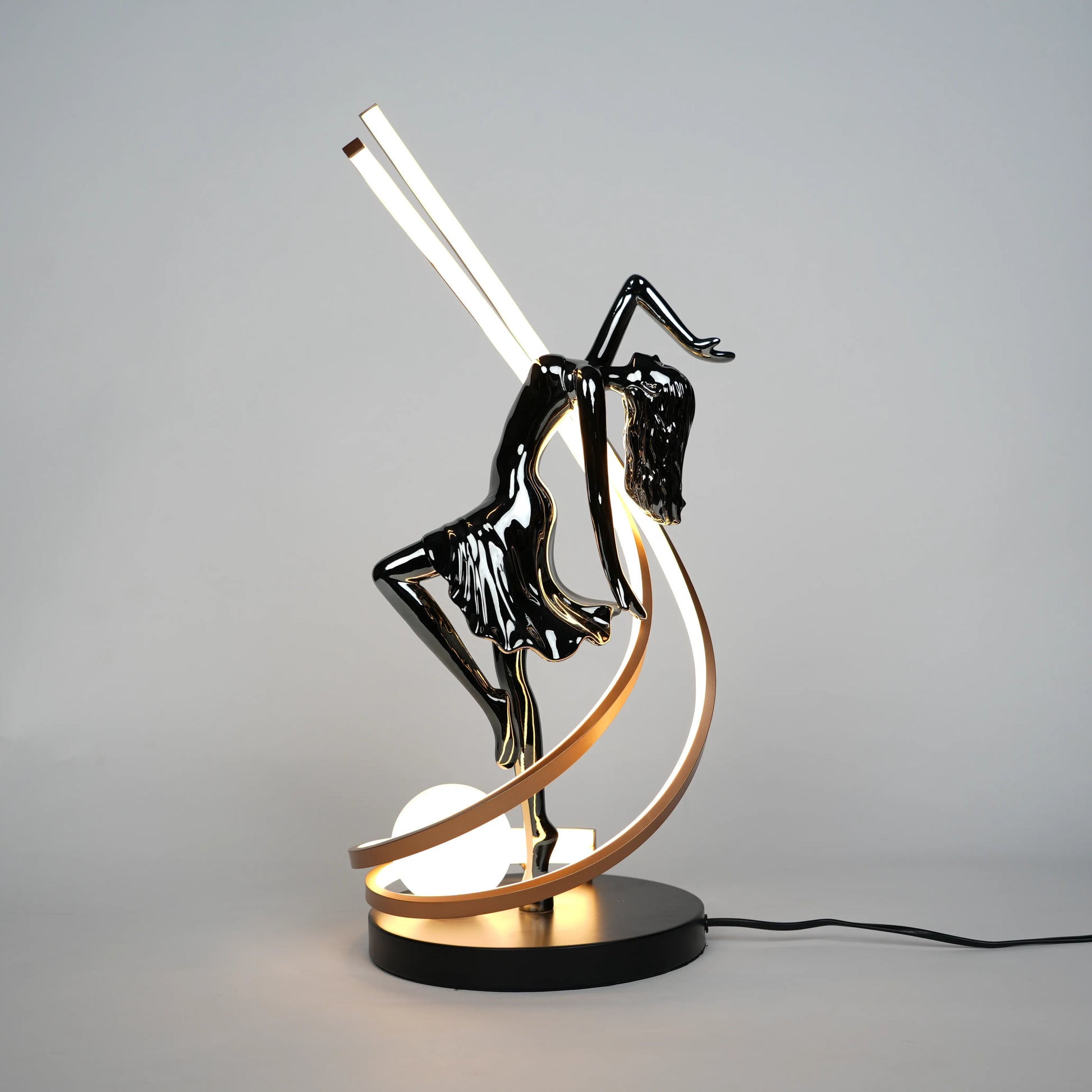 Small electroplated black sculpture lamp of a dancing woman with curved LED arcs and warm glowing orb.