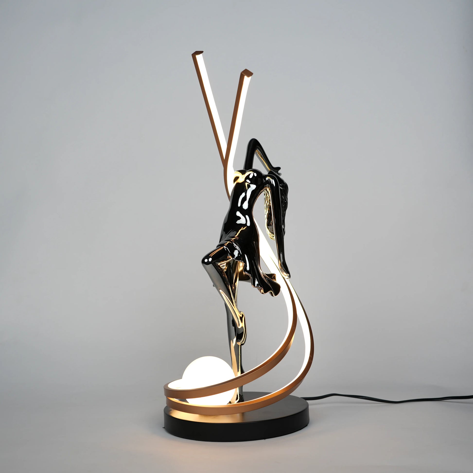 Small electroplated black sculpture lamp of a dancing woman with curved LED arcs and warm glowing orb.