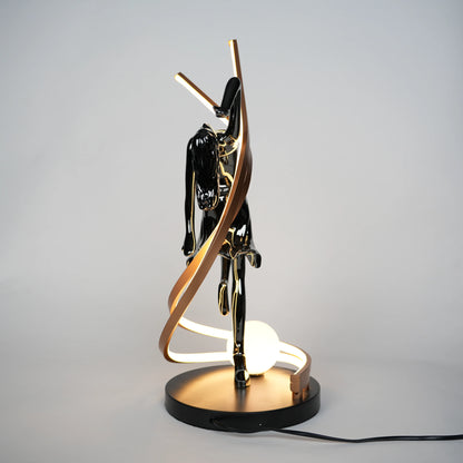 Small electroplated black sculpture lamp of a dancing woman with curved LED arcs and warm glowing orb.