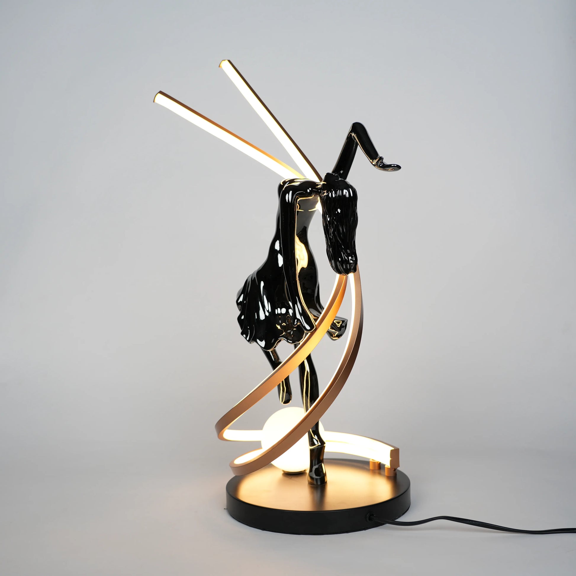 Small electroplated black sculpture lamp of a dancing woman with curved LED arcs and warm glowing orb.