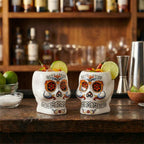 Two skull-shaped cocktail glasses with colorful designs on a bar counter.