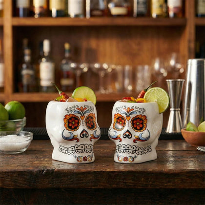 Two skull-shaped cocktail glasses with colorful designs on a bar counter.