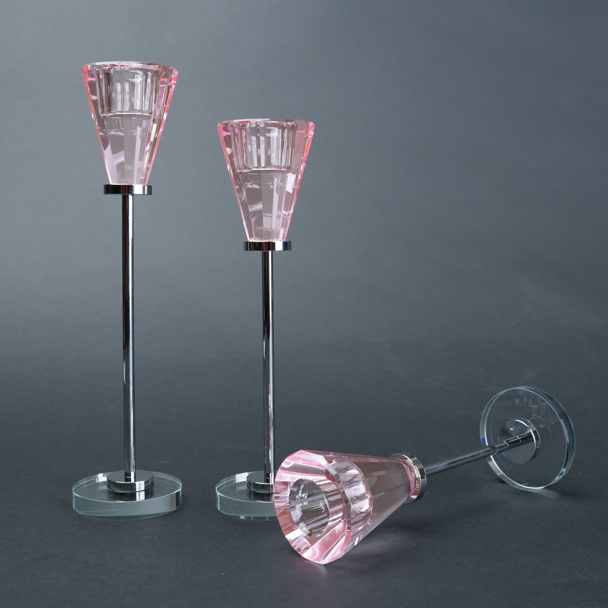 Two pink glass candle holders on stands with a third one lying on a dark gray background