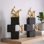 Two gold sculpture figures on stacked black and white blocks against a light gray background