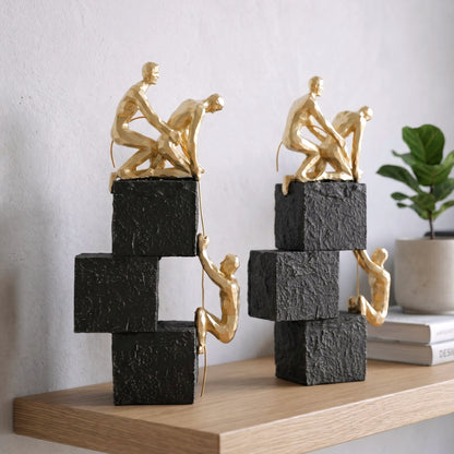 Two gold sculpture figures on stacked black and white blocks against a light gray background