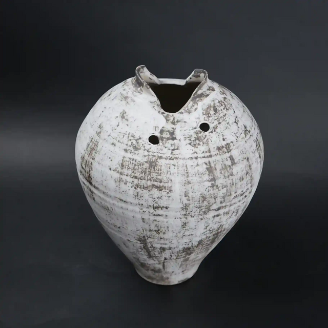 White distressed ceramic vase with rounded body and narrow base, featuring a textured rustic finish, dual side openings, and an uneven top rim, ideal for modern home decor and artistic styling.