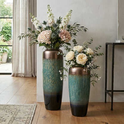 Two tall ceramic vases with floral arrangements on a wooden floor.