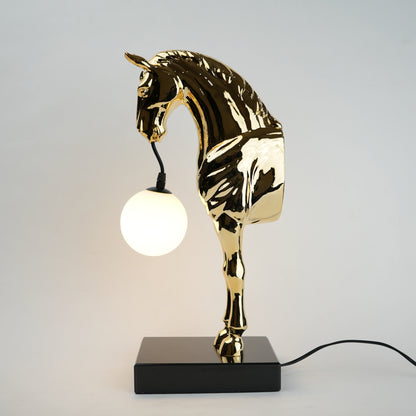 Golden horse table lamp with a sculpted horse base holding a frosted spherical globe light, featuring a smooth metallic finish and a structured base designed for soft ambient lighting and decorative use.