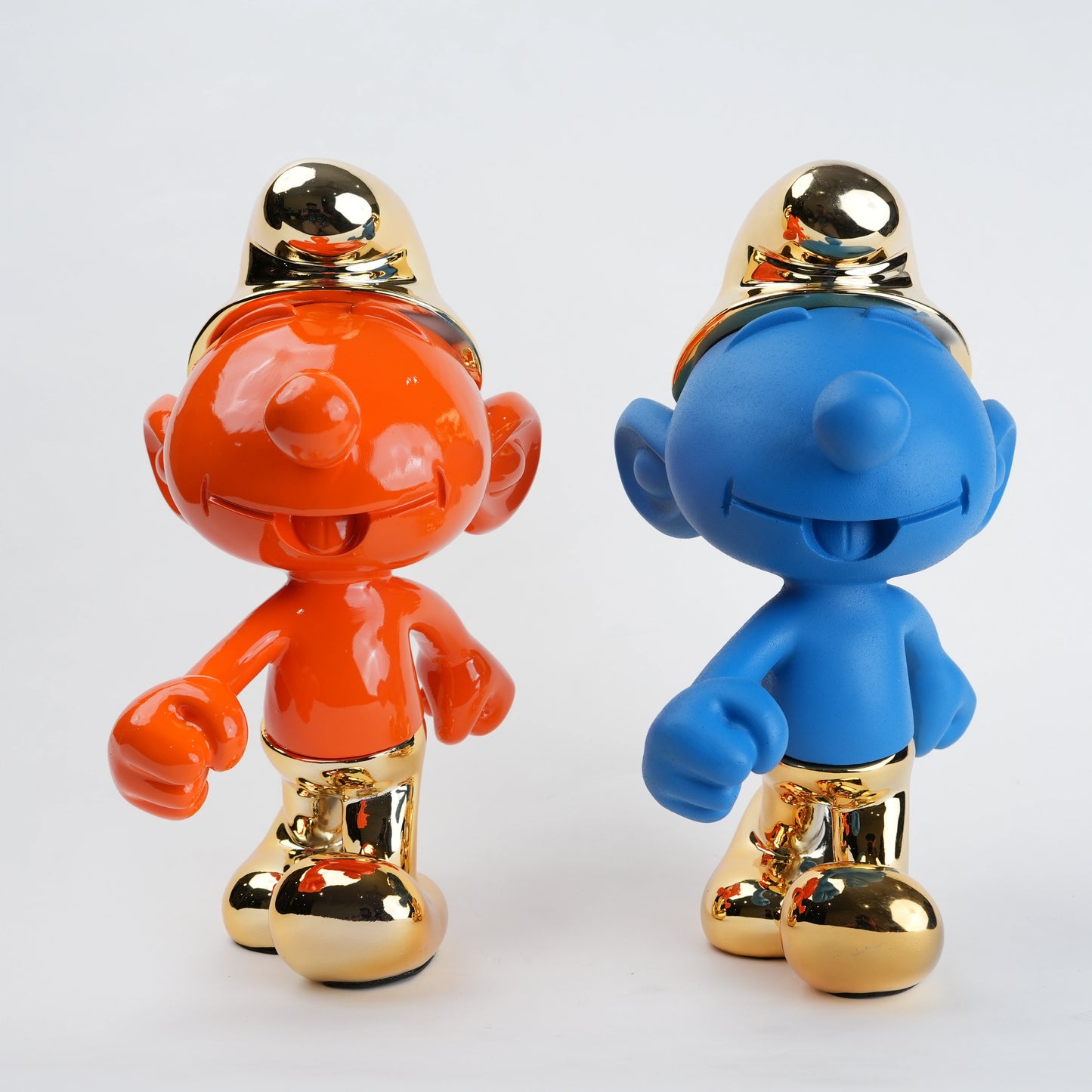 Two Smurf figurines, one orange and one blue, with gold hats and shoes on a light gray background.