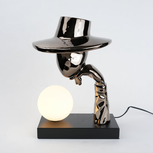 Black electroplated table lamp featuring a sculptural curved human-like form with a wide hat-shaped top and a glowing spherical light placed on a rectangular base, designed with a smooth reflective metallic surface.
