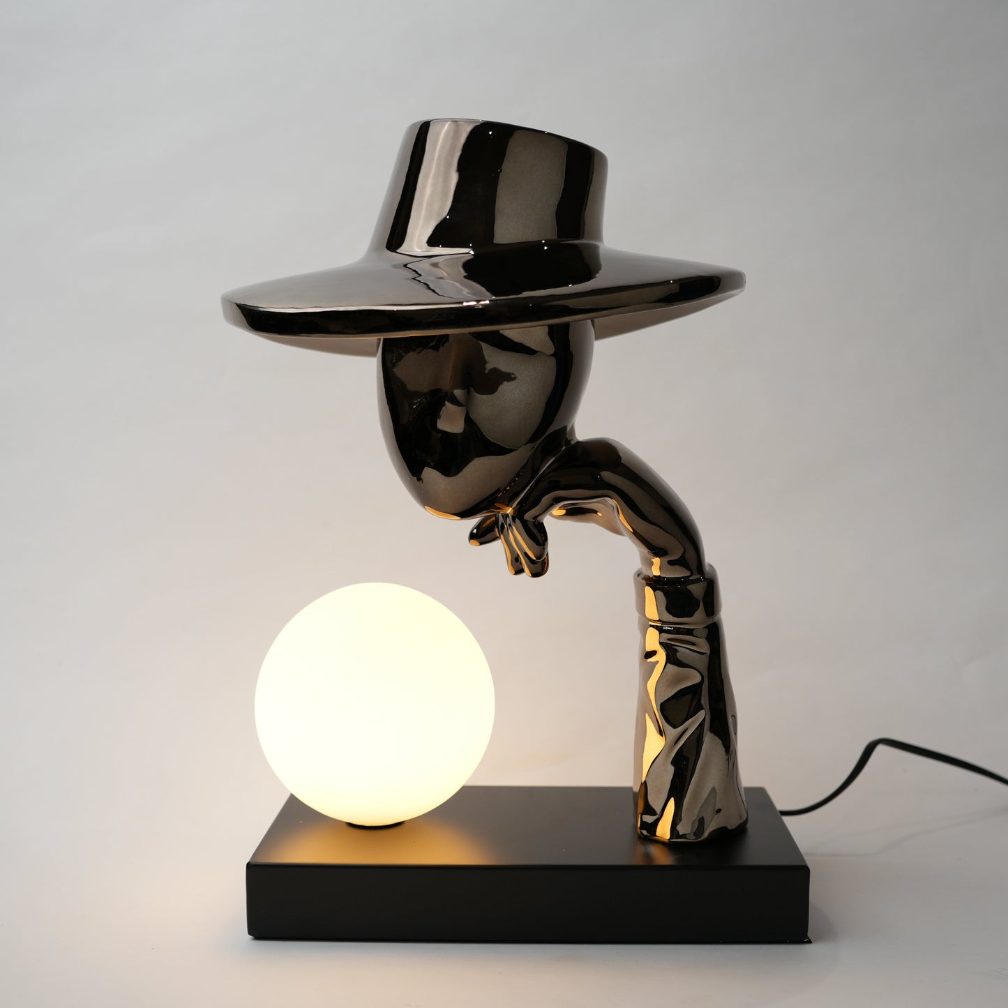 Black electroplated table lamp featuring a sculptural curved human-like form with a wide hat-shaped top and a glowing spherical light placed on a rectangular base, designed with a smooth reflective metallic surface.