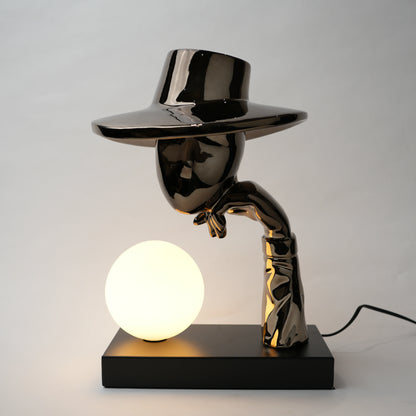 Black electroplated table lamp featuring a sculptural curved human-like form with a wide hat-shaped top and a glowing spherical light placed on a rectangular base, designed with a smooth reflective metallic surface.