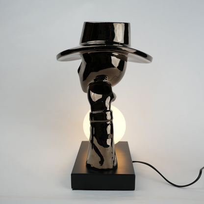 Black electroplated table lamp featuring a sculptural curved human-like form with a wide hat-shaped top and a glowing spherical light placed on a rectangular base, designed with a smooth reflective metallic surface.
