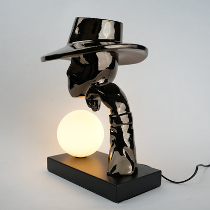 Black electroplated table lamp featuring a sculptural curved human-like form with a wide hat-shaped top and a glowing spherical light placed on a rectangular base, designed with a smooth reflective metallic surface.