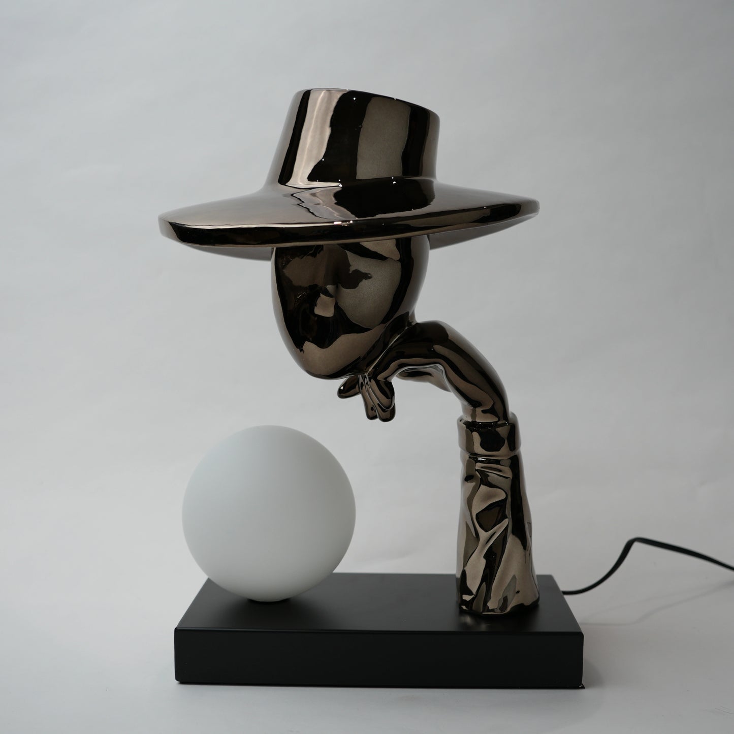 Black electroplated table lamp featuring a sculptural curved human-like form with a wide hat-shaped top and a glowing spherical light placed on a rectangular base, designed with a smooth reflective metallic surface.