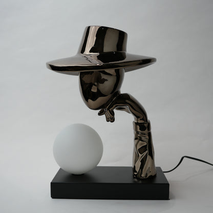 Black electroplated table lamp featuring a sculptural curved human-like form with a wide hat-shaped top and a glowing spherical light placed on a rectangular base, designed with a smooth reflective metallic surface.