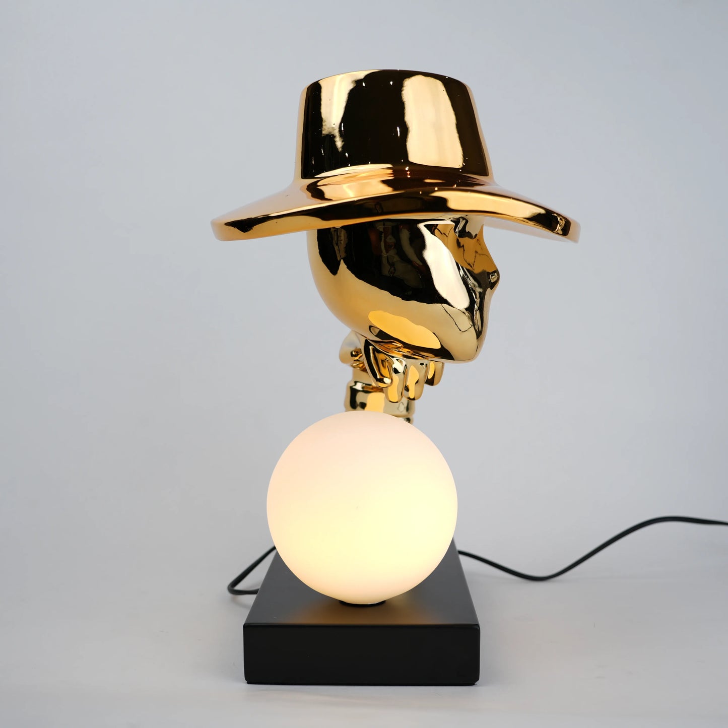 Gold electroplated table lamp featuring a sculptural curved human-like form with a wide hat-shaped top and a glowing spherical light on a rectangular base, designed with a smooth reflective metallic surface.