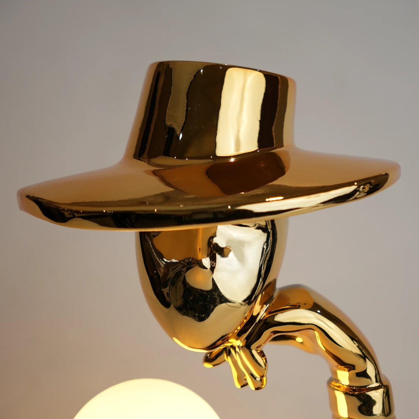 Gold electroplated table lamp featuring a sculptural curved human-like form with a wide hat-shaped top and a glowing spherical light on a rectangular base, designed with a smooth reflective metallic surface.