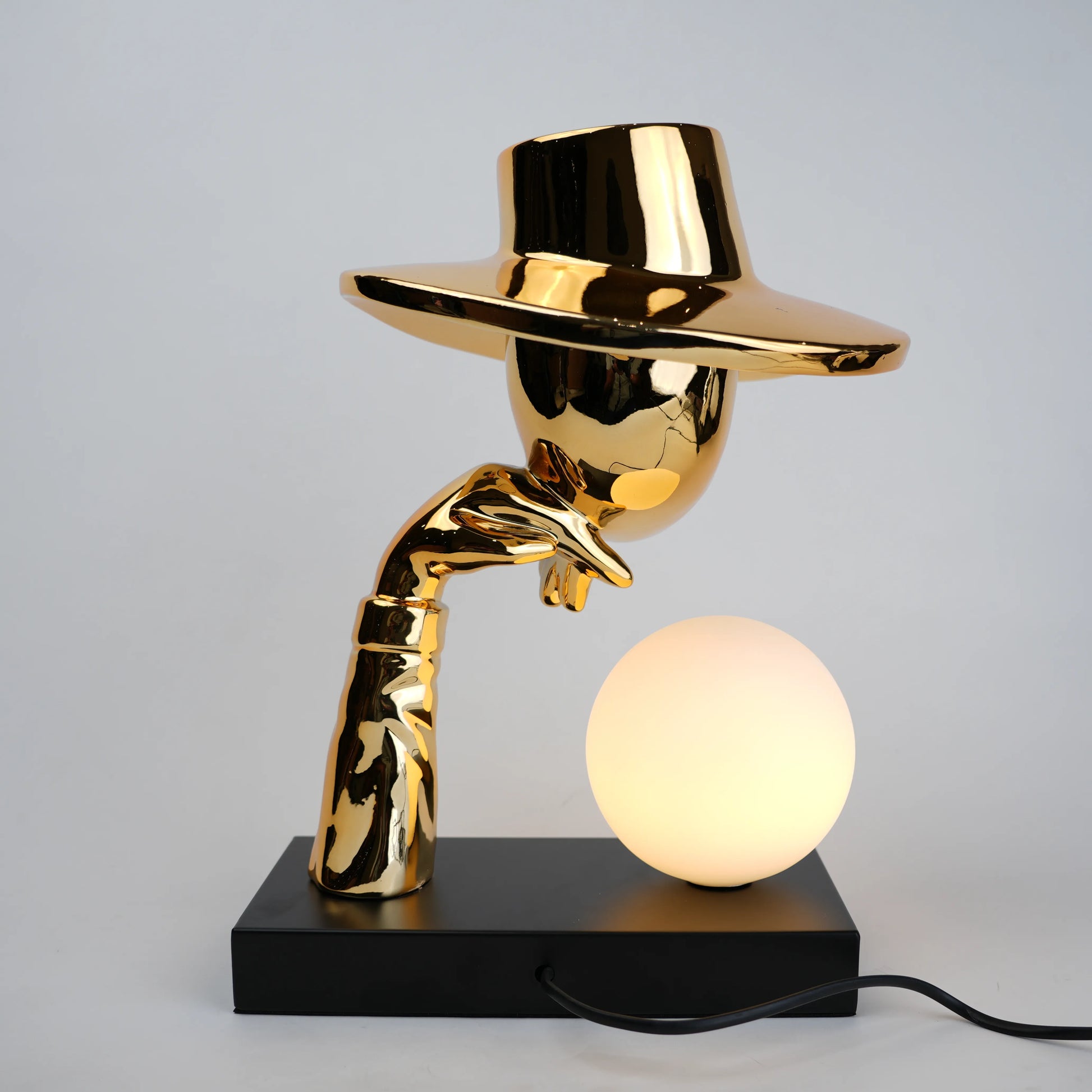 Gold electroplated table lamp featuring a sculptural curved human-like form with a wide hat-shaped top and a glowing spherical light on a rectangular base, designed with a smooth reflective metallic surface.