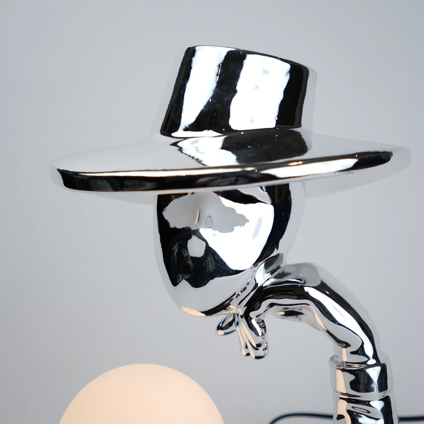 Silver electroplated table lamp featuring a sculptural curved human-like form with a wide hat-shaped top and a glowing spherical light on a rectangular base, finished with a smooth reflective metallic surface.