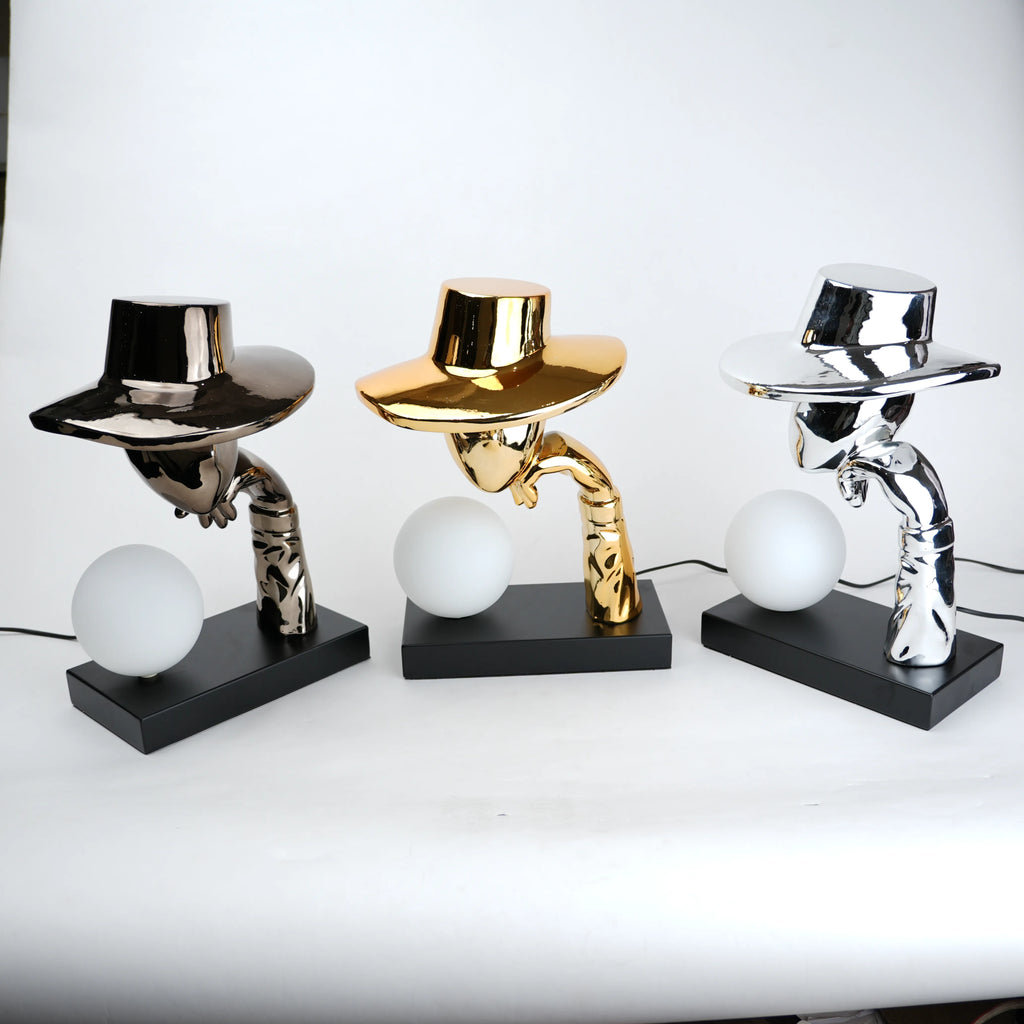 Silver electroplated table lamp featuring a sculptural curved human-like form with a wide hat-shaped top and a glowing spherical light on a rectangular base, finished with a smooth reflective metallic surface.