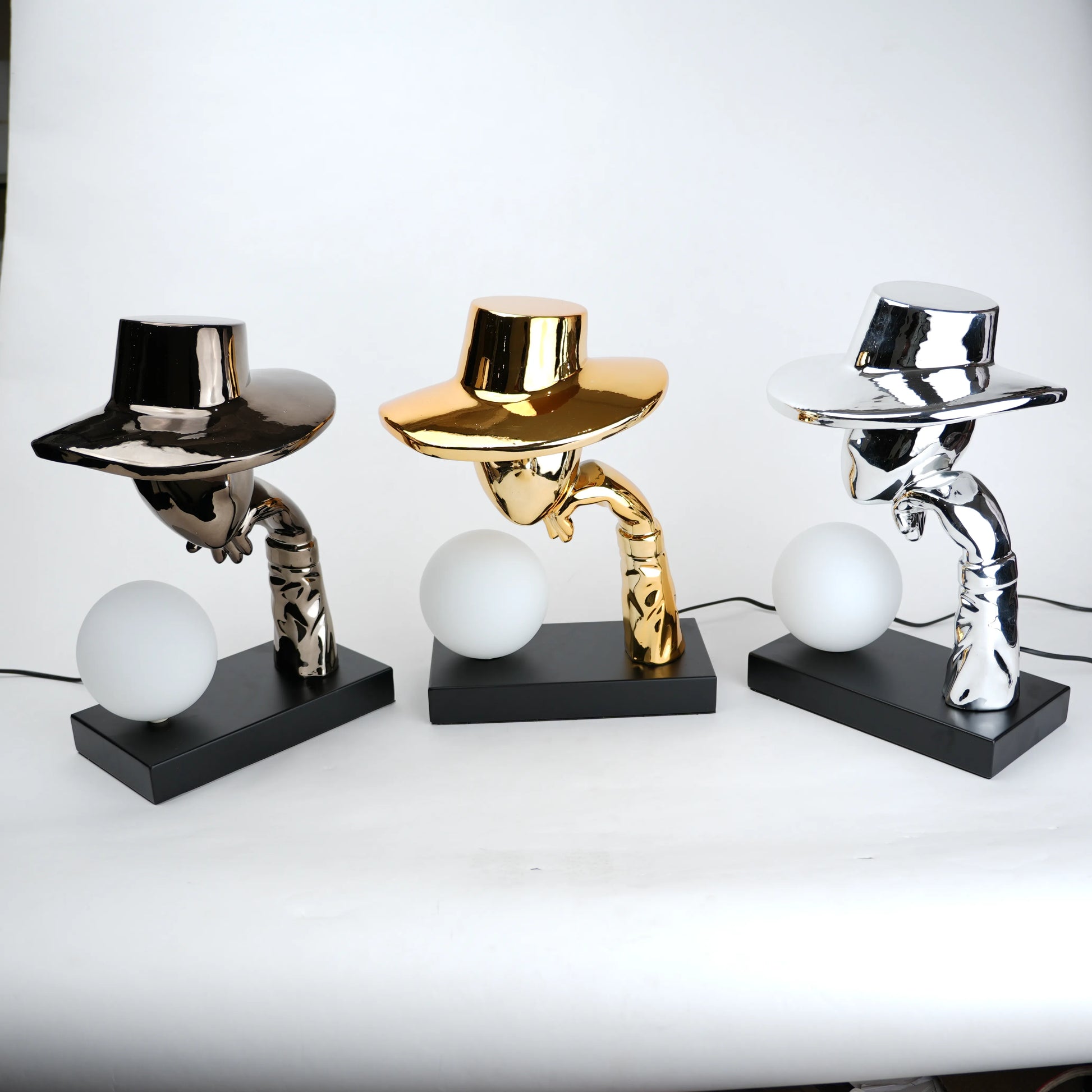 Silver electroplated table lamp featuring a sculptural curved human-like form with a wide hat-shaped top and a glowing spherical light on a rectangular base, finished with a smooth reflective metallic surface.