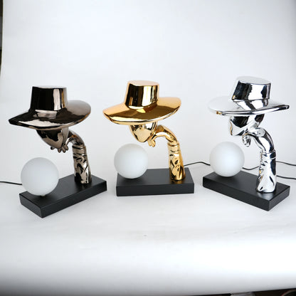 Silver electroplated table lamp featuring a sculptural curved human-like form with a wide hat-shaped top and a glowing spherical light on a rectangular base, finished with a smooth reflective metallic surface.