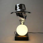 Silver electroplated table lamp featuring a sculptural curved human-like form with a wide hat-shaped top and a glowing spherical light on a rectangular base, finished with a smooth reflective metallic surface.