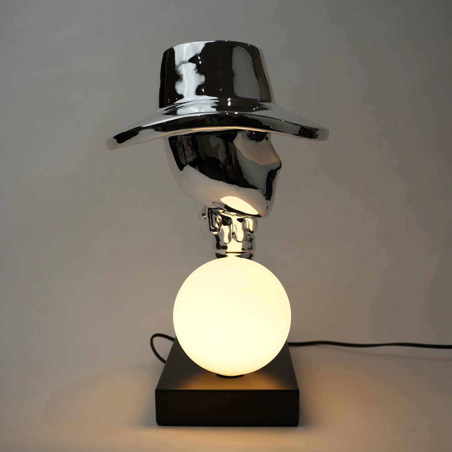 Silver electroplated table lamp featuring a sculptural curved human-like form with a wide hat-shaped top and a glowing spherical light on a rectangular base, finished with a smooth reflective metallic surface.