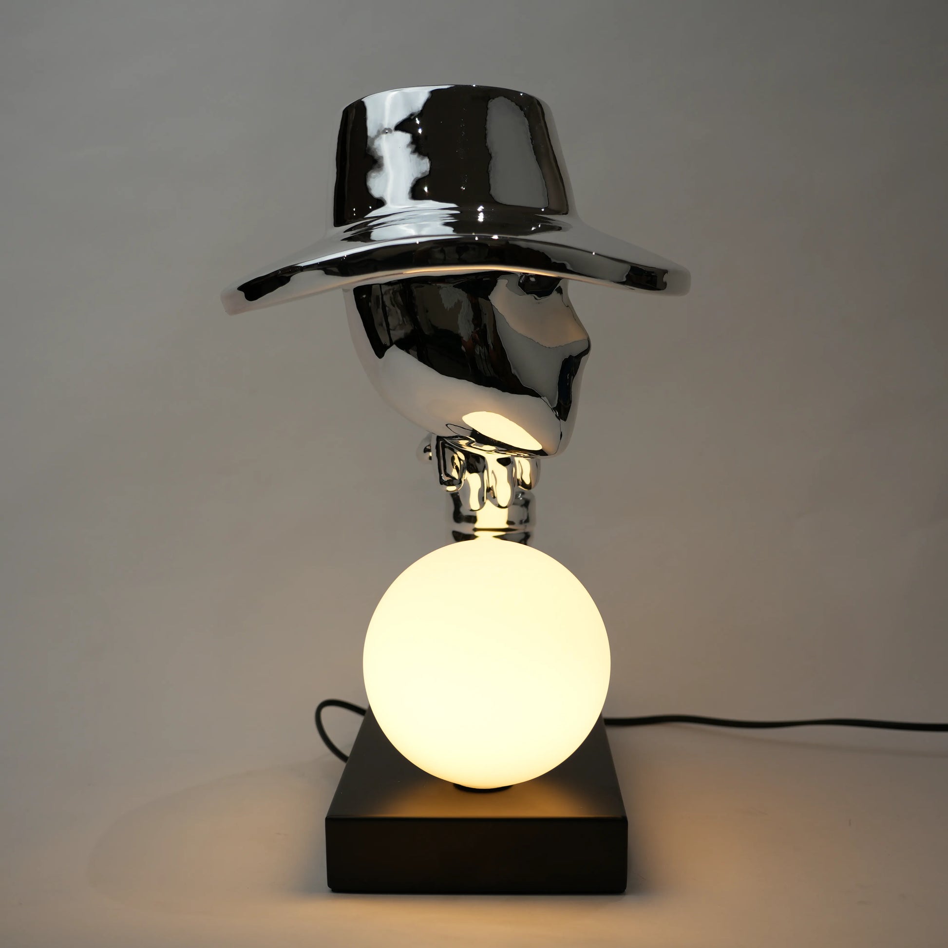 Silver electroplated table lamp featuring a sculptural curved human-like form with a wide hat-shaped top and a glowing spherical light on a rectangular base, finished with a smooth reflective metallic surface.