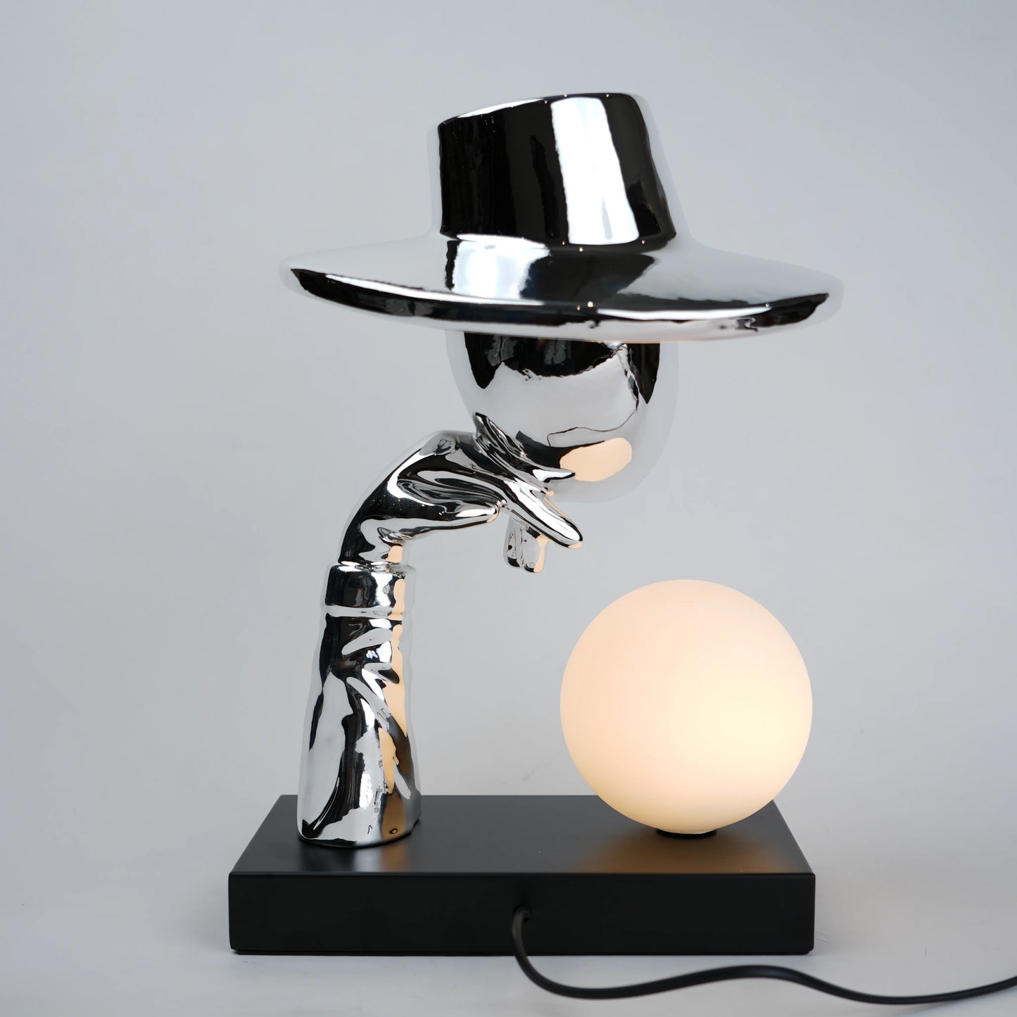 Silver electroplated table lamp featuring a sculptural curved human-like form with a wide hat-shaped top and a glowing spherical light on a rectangular base, finished with a smooth reflective metallic surface.