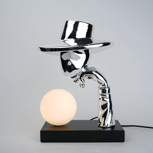 Silver electroplated table lamp featuring a sculptural curved human-like form with a wide hat-shaped top and a glowing spherical light on a rectangular base, finished with a smooth reflective metallic surface.