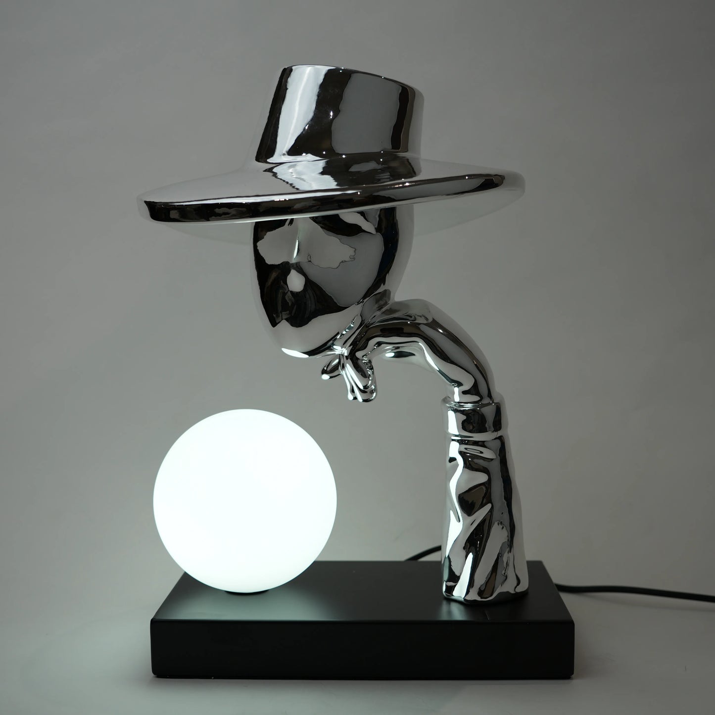 Metallic sculpture of a figure holding a glowing sphere on a black base against a gray background