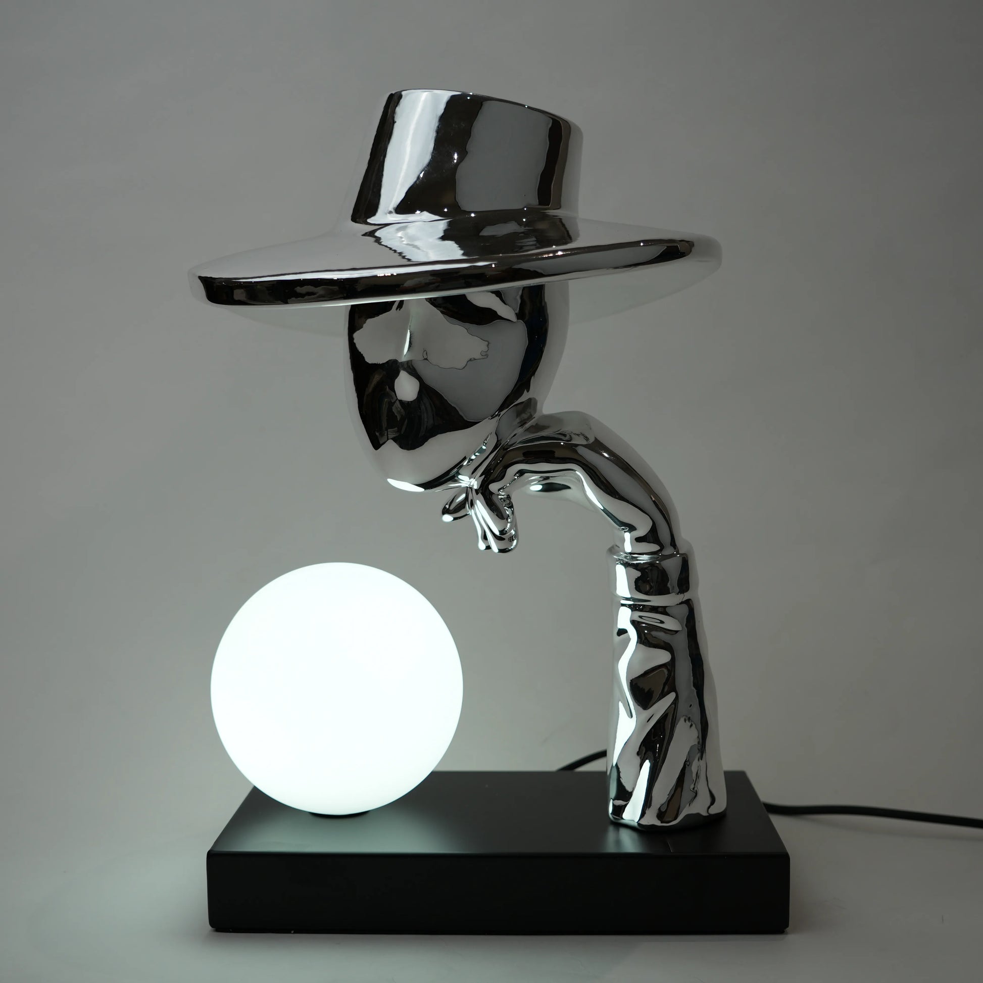 Metallic sculpture of a figure holding a glowing sphere on a black base against a gray background