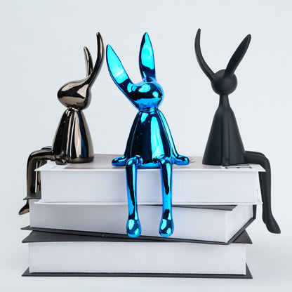 Three rabbit-shaped sculptures in black, blue, and silver on stacked books with a white background