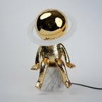 Golden human figurine table lamp with textured metallic finish sitting on a white marble base, featuring a reflective spherical head encircled by an illuminated LED halo ring emitting warm ambient light.