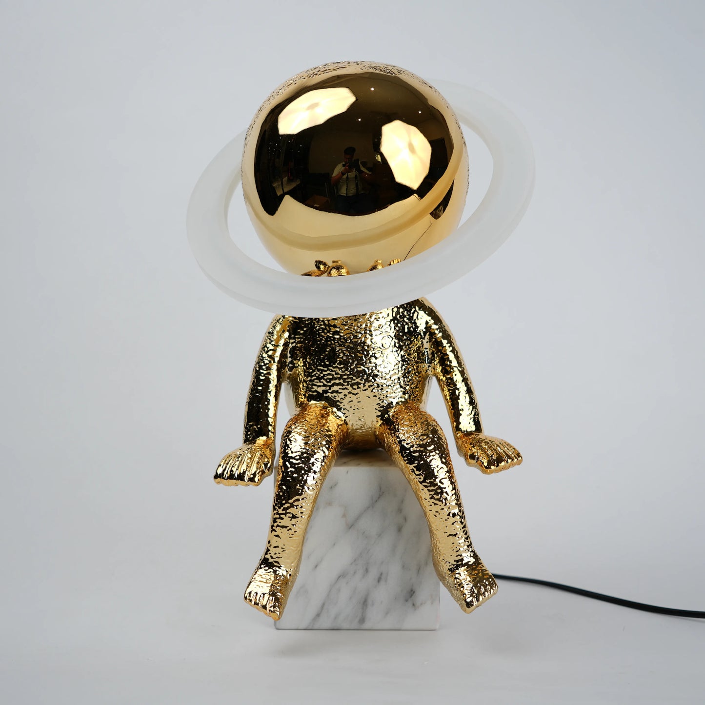 Golden human figurine table lamp with textured metallic finish sitting on a white marble base, featuring a reflective spherical head encircled by an illuminated LED halo ring emitting warm ambient light.