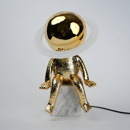 Golden human figurine table lamp with textured metallic finish sitting on a white marble base, featuring a reflective spherical head encircled by an illuminated LED halo ring emitting warm ambient light.