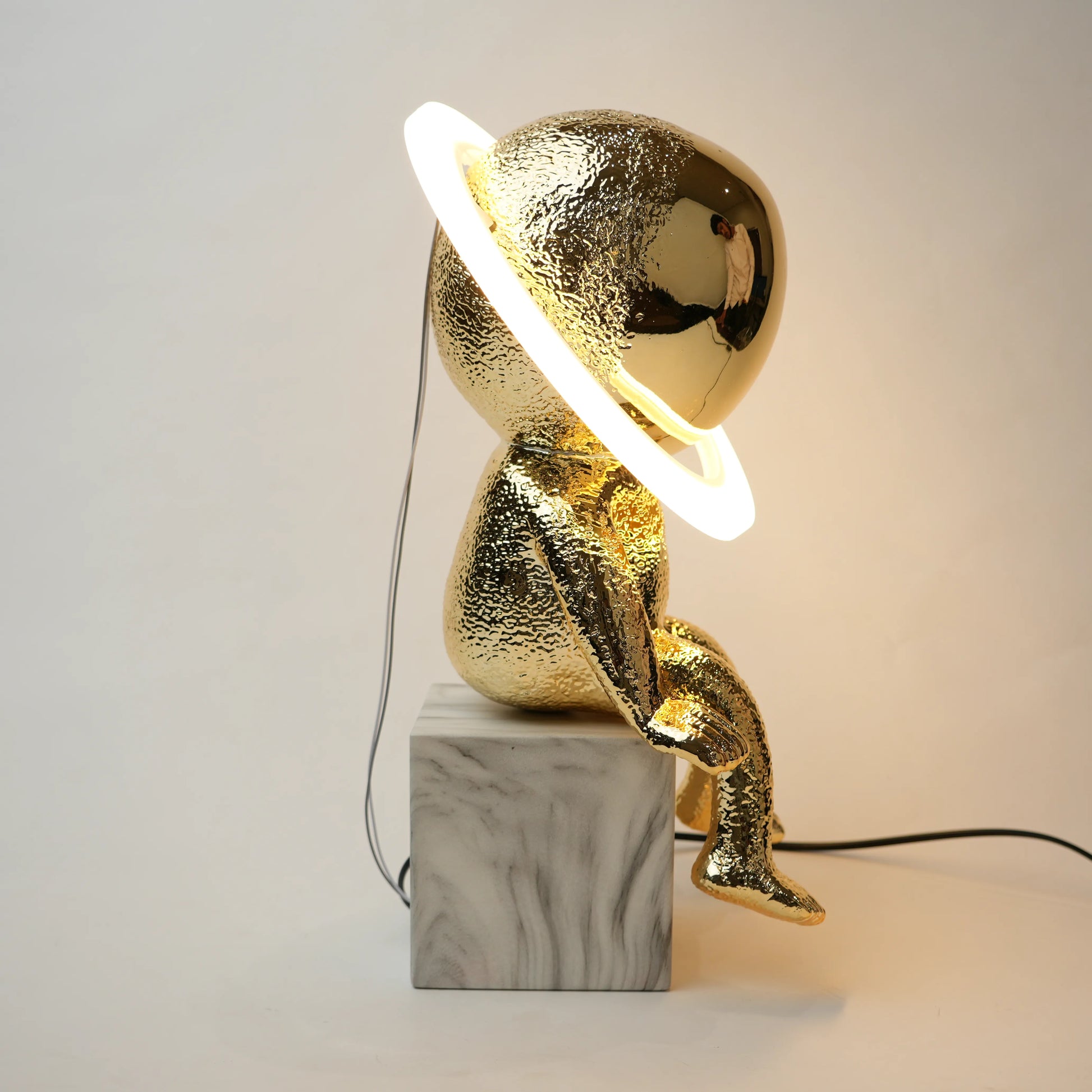 Golden human figurine table lamp with textured metallic finish sitting on a white marble base, featuring a reflective spherical head encircled by an illuminated LED halo ring emitting warm ambient light.