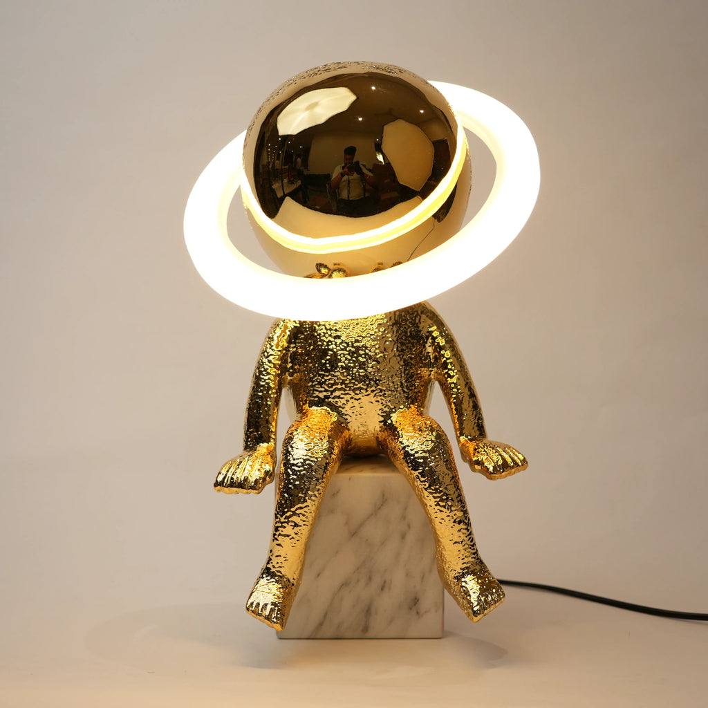 Golden human figurine table lamp with textured metallic finish sitting on a white marble base, featuring a reflective spherical head encircled by an illuminated LED halo ring emitting warm ambient light.