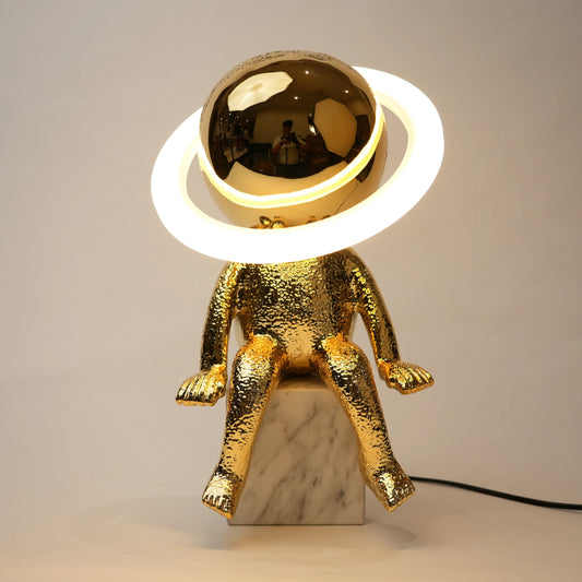 Golden human figurine table lamp with textured metallic finish sitting on a white marble base, featuring a reflective spherical head encircled by an illuminated LED halo ring emitting warm ambient light.