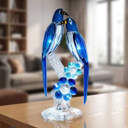 A hand holds a blue and clear crystal bird sculpture of two birdsa perched on a flower, against a plain light gray background.