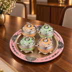 Four ornate lidded glass jars with silver-toned animal engravings and pastel orange and green interiors, arranged on a round pink and silver decorative tray against a black background.