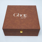 A square brown faux-leather gift box with a gold clasp and the brand name 'Ghar art' printed in white on the lid, set against a light gray background.