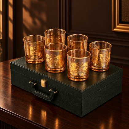 Six amber-tinted glass tumblers with gold floral etched patterns, arranged on top of a dark green textured presentation box with a gold clasp, against a white background.