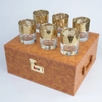 A set of six gold-patterned glass tumblers arranged on top of a brown wood-grain storage box with a gold combination lock and handle