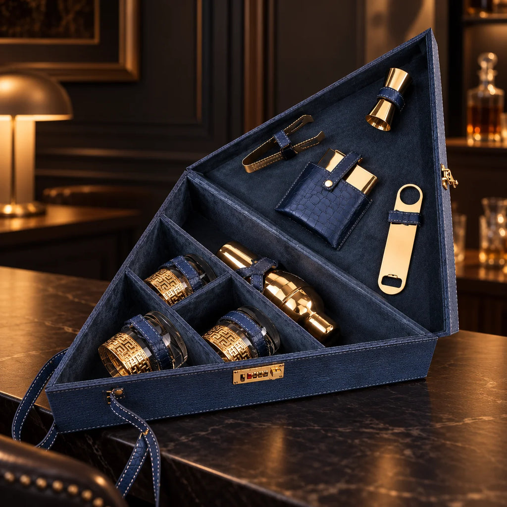 A dark gray wooden cocktail gift set organizer shown open, containing gold-toned metal bar tools and bottles with dark blue leather straps.