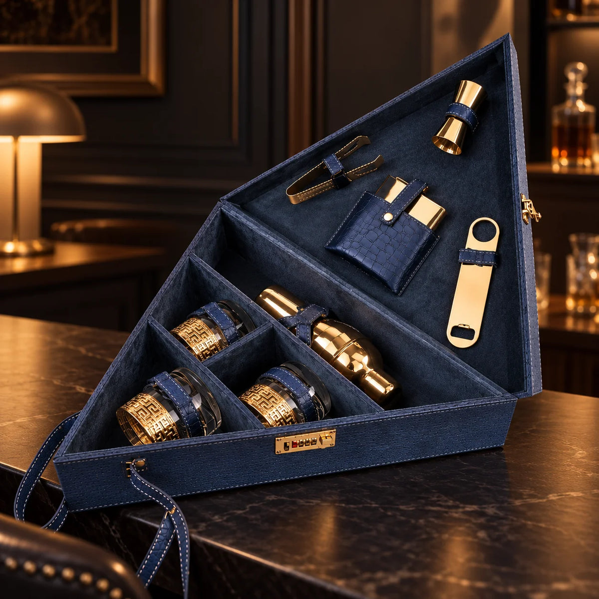 A dark gray wooden cocktail gift set organizer shown open, containing gold-toned metal bar tools and bottles with dark blue leather straps.