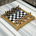 A close-up of ornate gold chess pieces arranged on a glossy black-and-white checkered board with a gold frame, photographed in a real indoor setting with a blurred sofa and pillow in the background.