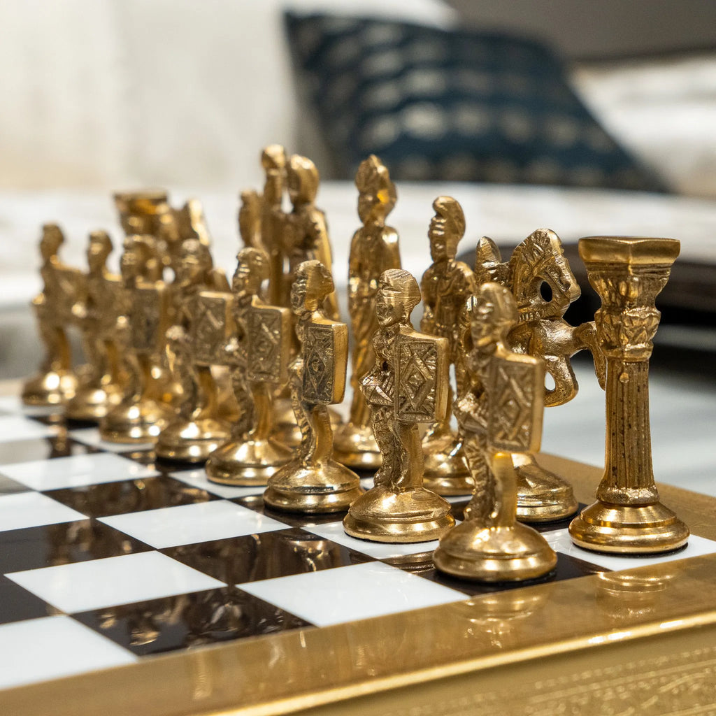 A close-up of ornate gold chess pieces arranged on a glossy black-and-white checkered board with a gold frame, photographed in a real indoor setting with a blurred sofa and pillow in the background.